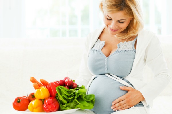 Cheerful beautiful pregnant woman sitting on the sofa with a bowl full of vegetables.